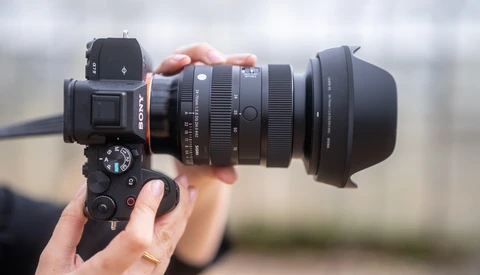Photographer holding a mirrorless camera with telephoto lens and hood attached outdoors.
