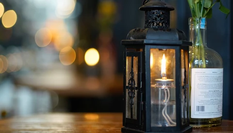 Black metal lantern with lit candle next to a wine bottle on wooden surface with bokeh lights in background.
