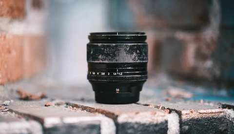 Black vintage camera lens with aperture markings positioned on weathered concrete surface with blurred brick background.