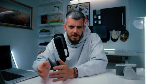 Man in gray hoodie demonstrating a handheld camera or recording device at a desk.