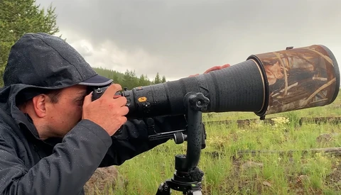 Photographer using a telephoto lens with camouflage barrel cover mounted on a tripod in an open field.