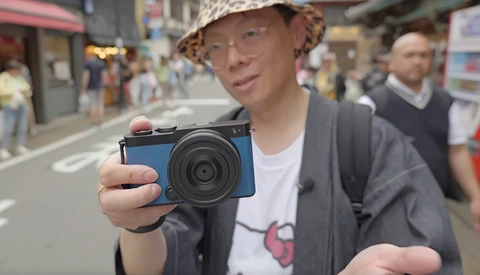 Photographer holding a blue compact digital camera while standing in an urban street.