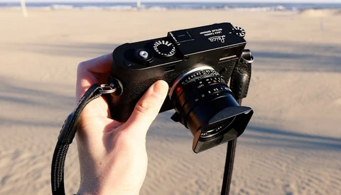Hand holding a rangefinder camera with attached lens against a beach background.