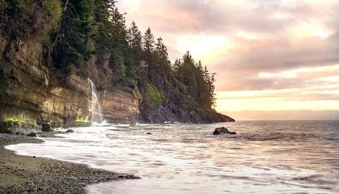 Coastal cliff with evergreen forest and waterfall cascading onto rocky beach at sunset.