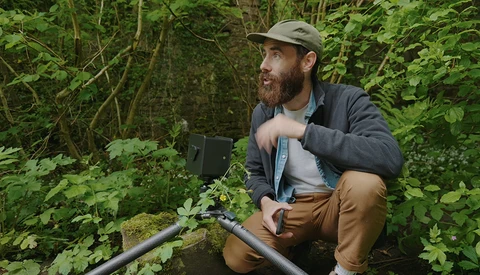 Cinematographer crouching beside a creek in dense forest vegetation, holding audio recording equipment.