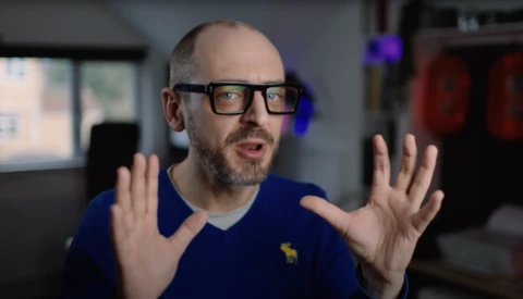 Man with glasses wearing navy blue shirt gestures expressively while speaking in studio setting.