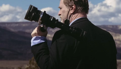 Photographer in dark jacket holding a telephoto lens to his eye, photographing a vast canyon landscape under blue sky.