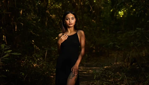 Woman in black dress standing in darkened forest with warm golden lighting filtering through trees.