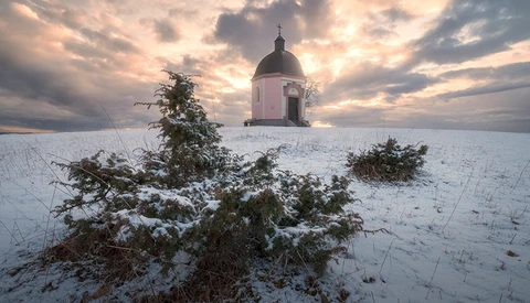 Snow-covered landscape with a domed chapel and bare shrubs under dramatic cloudy sky.