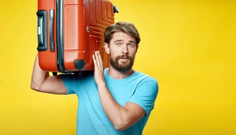 Bearded man in light blue t-shirt carrying red suitcase on shoulder against yellow background.