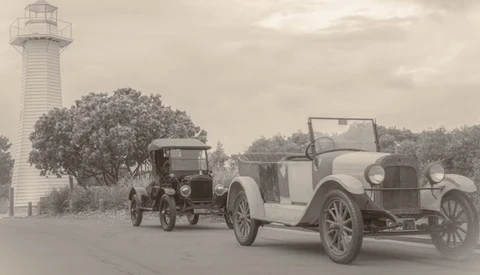 Two vintage automobiles parked near a lighthouse under overcast sky.