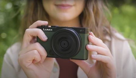 Woman holding a black Lumix compact camera outdoors with blurred green background.