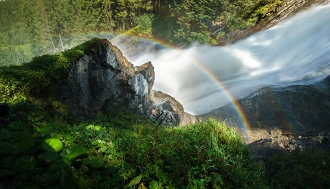 Waterfall with rainbow mist against forested mountainside and rocky outcrop.