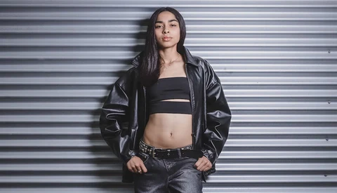 Woman in black leather jacket and crop top posing against corrugated metal wall.