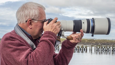 Older photographer holding a telephoto lens up to his eye while standing at a railing overlooking water.