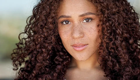 Portrait of a woman with long curly brown hair and freckles looking directly at camera.