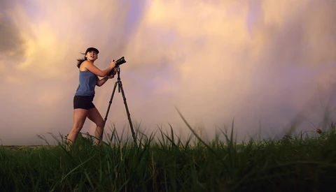 Woman operating a telescope on a tripod in a grassy field at golden hour with dramatic clouds.