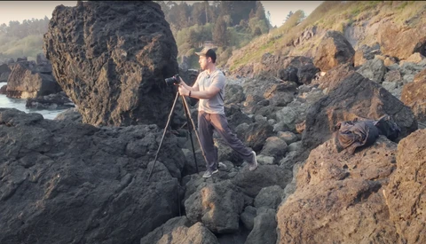 Photographer on tripod positioning among rocky coastal formations with water and forested hills in background.