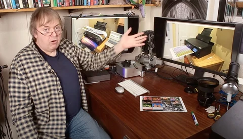 Photographer gestures toward camera equipment displayed on a computer monitor in a home studio setup.