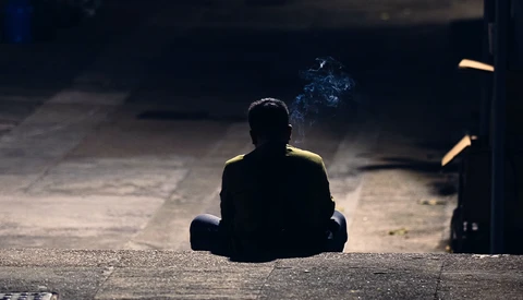 Silhouetted figure sitting cross-legged on concrete ground under dramatic directional lighting.