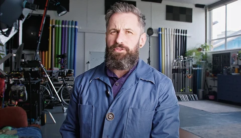 Bearded man in blue shirt standing in professional photography studio with lighting equipment and grip gear visible in background.
