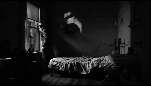A figure suspended above a bed in a dramatically lit black and white interior scene.