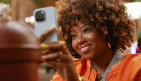 Woman in orange jacket holding white smartphone, smiling at camera in warm indoor lighting.