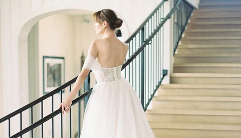 Bride in white off-shoulder gown descending staircase, hand on railing, viewed from behind.