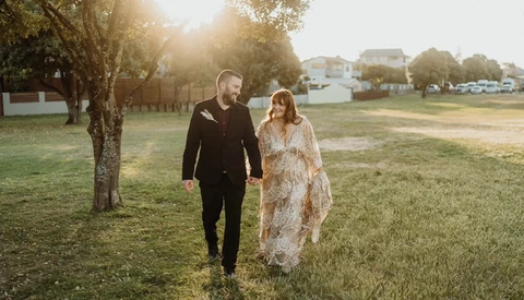 Couple walking hand-in-hand through a grassy field at golden hour with trees and houses in the background.