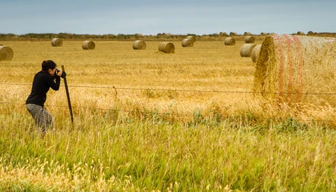Photographer with camera on tripod capturing hay bales in rural field.