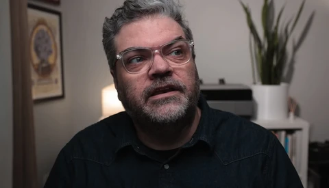 Man with gray hair and beard wearing glasses and dark button-up shirt in an office setting.