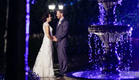 Bride and groom holding hands at night under purple ambient lighting and illuminated trees.