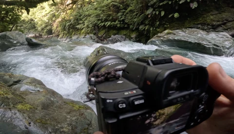 Photographer holding a mirrorless camera with wide-angle lens pointed at flowing stream surrounded by rocks and forest.