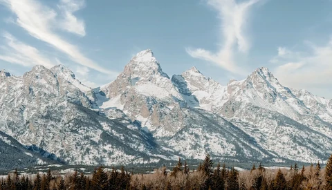 Snow-covered mountain range with forested foothills under blue sky with wispy clouds.