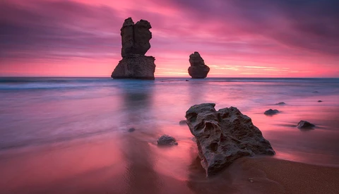 Rocky sea stacks silhouetted against a vibrant pink and purple sunset reflected in calm shallow water.