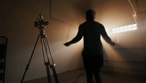 Silhouetted figure gesturing expressively toward a camera on tripod in a bright, hazy studio space.