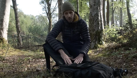 Man crouching beside backpack in forest clearing surrounded by tall trees.