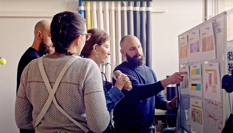 A bearded man in dark clothing presents design mockups on a wall to a group of four colleagues in an indoor studio or office space.