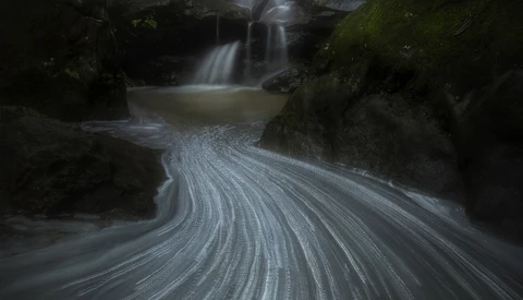 Long-exposure photograph of a flowing stream with silky water patterns curving around moss-covered rocks and a waterfall in the background.