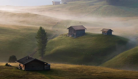 Misty alpine valley with scattered wooden chalets nestled among rolling green hills at golden hour.