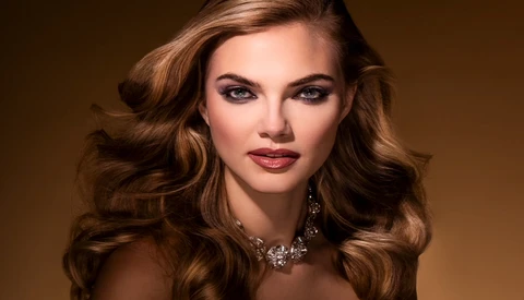 Woman with voluminous wavy brunette hair and dark red lipstick wearing a decorative statement necklace against a warm brown background.