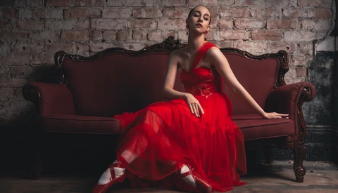 Woman in a red tulle gown seated on a burgundy velvet sofa against an exposed brick wall.