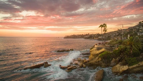 Rocky coastal cliff at sunset with palm trees and golden light reflecting on calm ocean waters.