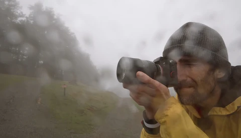 Photographer in yellow jacket and beanie shooting in heavy fog with binoculars-style camera positioning.