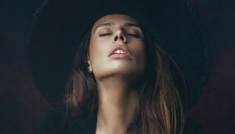 Woman with eyes closed wearing a black wide-brimmed hat against a dark background.