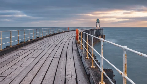 Wooden pier with white railings extending toward a lighthouse under dramatic cloudy sky at twilight.