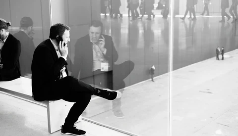 Businessman in dark suit sitting on white bench against glass window, speaking on phone while observing busy corridor reflection.