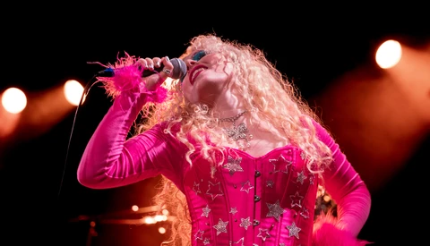 Performer in magenta sequined outfit and blonde curled wig singing into microphone on stage.