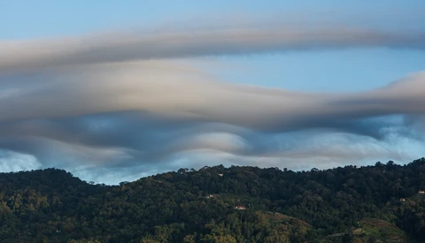 Dramatic lenticular clouds layered over a forested mountain landscape beneath blue sky.