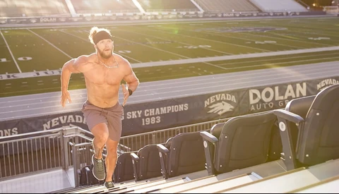 Shirtless runner in motion on a stadium press box with field and signage visible behind.